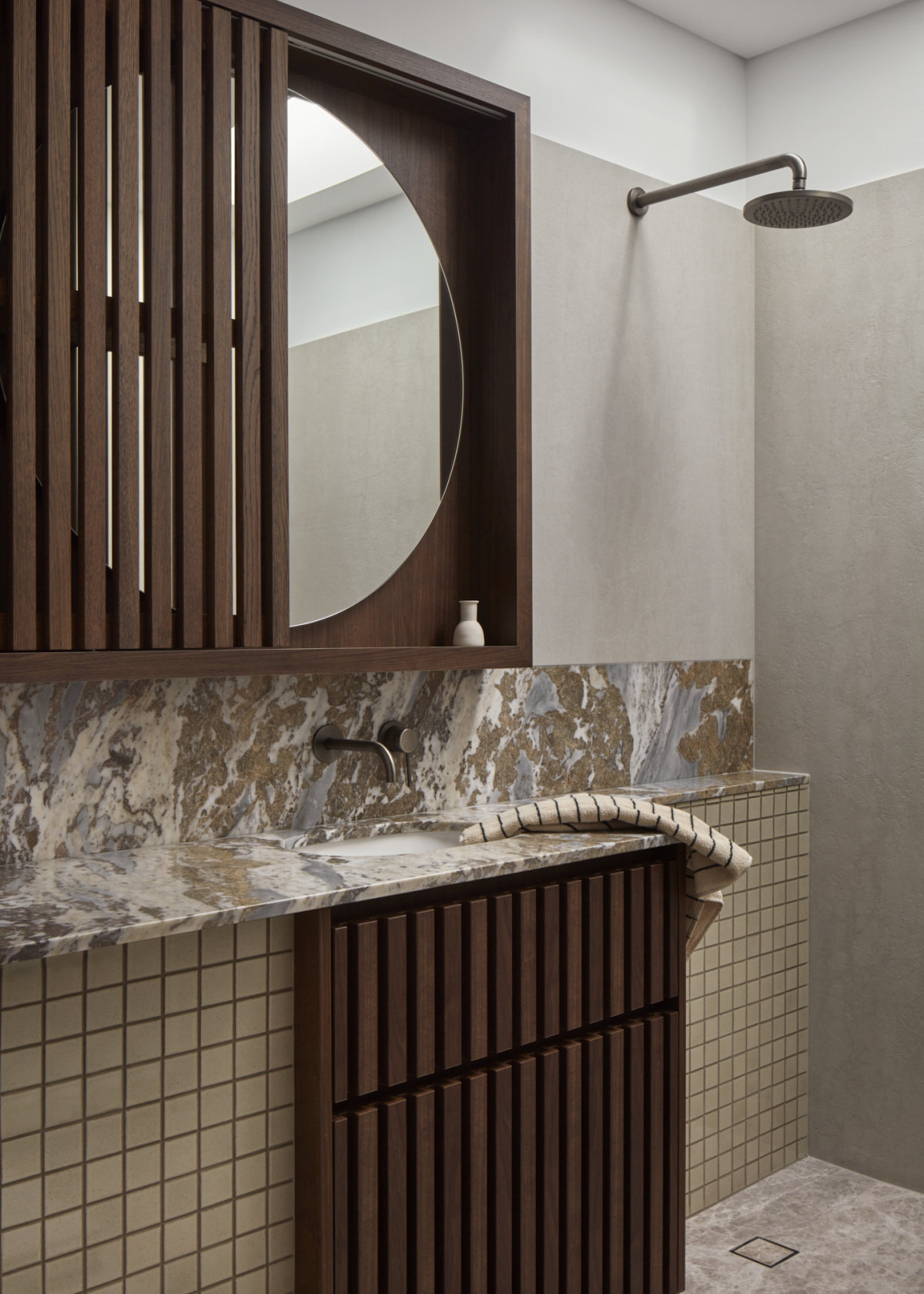 Bathroom vanity and shower area. Material like dark wood, light concrete, small beige tiles, and tri-colour patterned marble are used and arranged in balance.