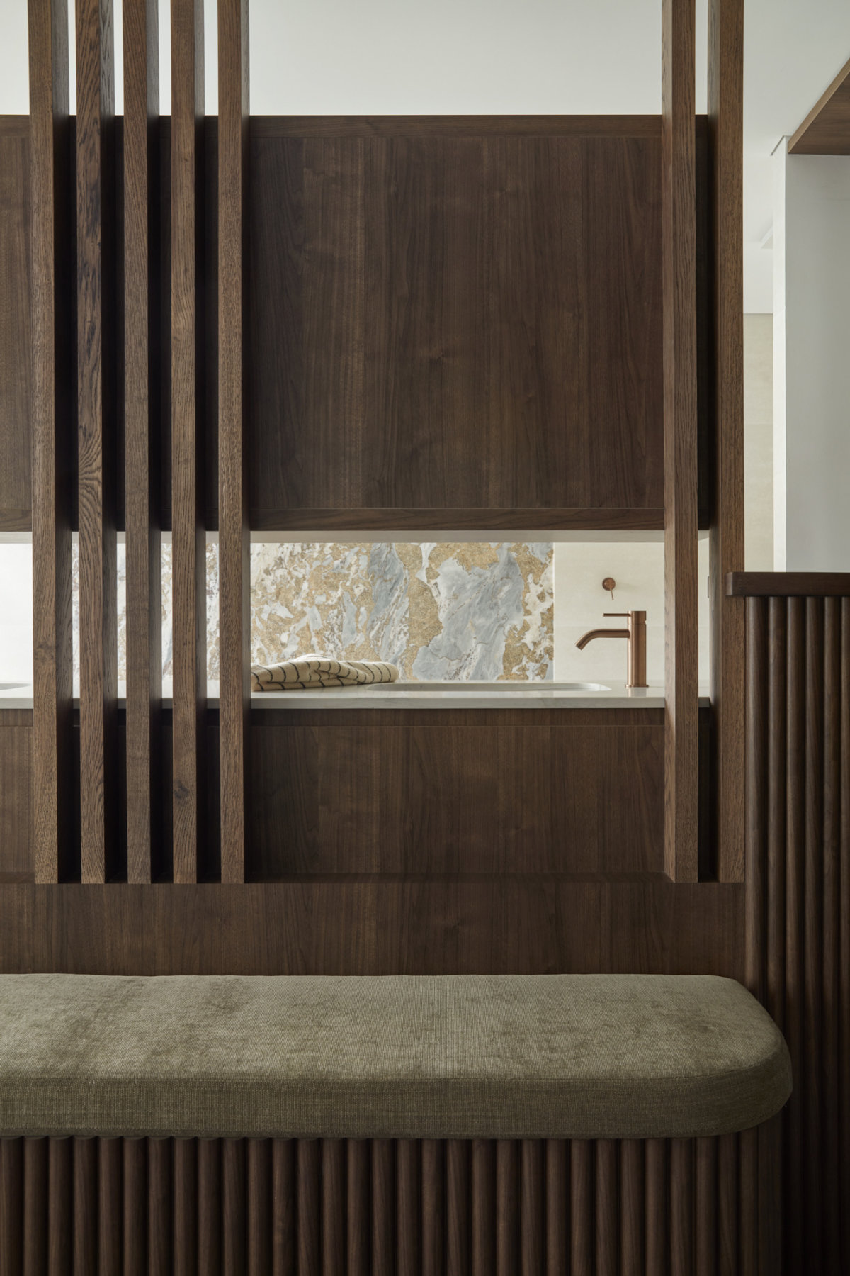 A straight-on view through the wooden slats partioning the bedroom and ensuite. The wall opposite is made of blue and brown patterned marble. In the foreground is a wide wooden bench with an olive cushioned top.